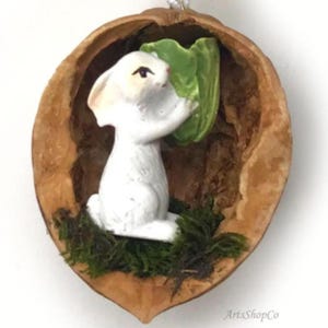 May include: A handcrafted ornament featuring a white bunny figurine holding a green leaf inside a walnut shell. The bunny sits on a bed of green moss. The shell is brown with a natural texture. A small silver string is attached for hanging.