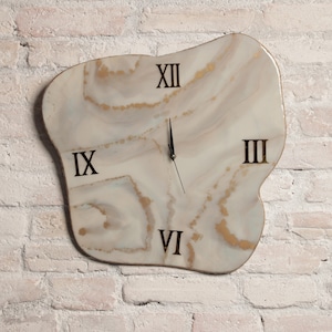 Puede incluir: Un reloj de resina blanco y dorado con números romanos. El reloj tiene una forma orgánica única y está montado en una pared de ladrillo blanco.