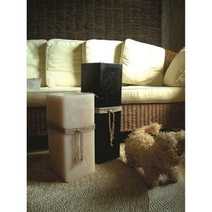 May include: Two square pillar candles, one white and one black, with rope tied around them. The candles are sitting on a woven rug in front of a wicker sofa.