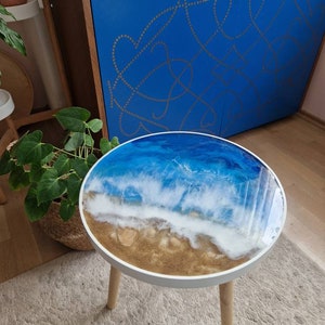 Può includere: Un tavolo rotondo in legno con un piano bianco e tre gambe in legno. Il piano del tavolo presenta un design in resina di onde oceaniche che si infrangono su una spiaggia sabbiosa.