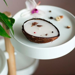 Peut inclure: Une bougie en forme de noix de coco blanche avec des accents bruns et des fleurs séchées sur un support à étages blanc.