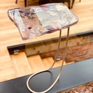 Könnte beinhalten: Ein kleiner, transparenter rechteckiger Tisch mit einem goldenen Metallgestell. Die Tischplatte besteht aus Harz und enthält gepresste Blumen und Blätter.