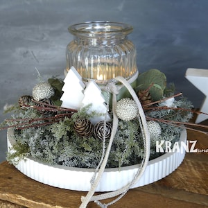 Könnte beinhalten: Festlicher Tischschmuck mit einem transparenten Glas-Kerzenhalter, umgeben von Grün, Tannenzapfen und weißen Dekorationselementen. Die Anordnung umfasst kleine weiße Holzbäume und silberne Ornamente.