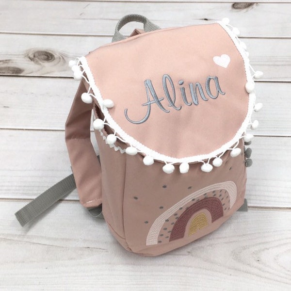 May include: A pink children's backpack with a white pom-pom trim and a rainbow embroidered on the front. The backpack has the name "Alina" embroidered on the flap.