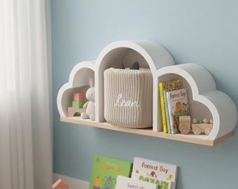 Könnte beinhalten: Ein weißes, wolkenförmiges Regal mit hellbraunem Holzsockel, das Bücher, Spielzeug und eine beige Aufbewahrungsbox mit dem Namen "Lean" enthält. Zusätzliche Bücher und Holzspielzeug liegen auf dem Boden, was ein spielerisches Thema für das Kinderzimmer schafft.