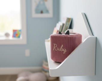 Könnte beinhalten: Ein weißes Wandregal hält eine rosa Aufbewahrungsbox mit schwarzen Katzenohren und dem Namen "Ruby" darauf gestickt. Das Regal befindet sich an einer hellblauen Wand, mit Büchern und einem Fenster.