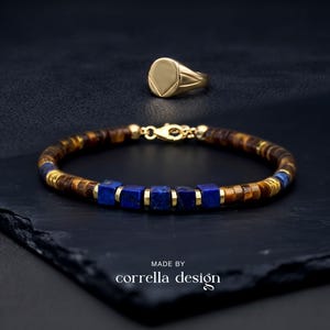 Könnte beinhalten: Ein Armband mit braunen Tigerauge-Scheiben, quadratischen blauen Lapislazuli-Perlen und goldfarbenen Zwischenelementen mit Karabinerverschluss, begleitet von einem goldfarbenen Siegelring. Der Text MADE BY corrella design ist unten sichtbar.