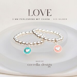 Könnte beinhalten: Zwei Perlenringe aus 925er Sterlingsilber, jeweils mit einem kleinen runden Anhänger. Ein Anhänger ist türkis mit einem silbernen Herz, der andere ist hellrosa mit einem silbernen Herz. Der Text LOVE und 3 MM PERLENRING MIT CHARM - 925 SILBER ist sichtbar.