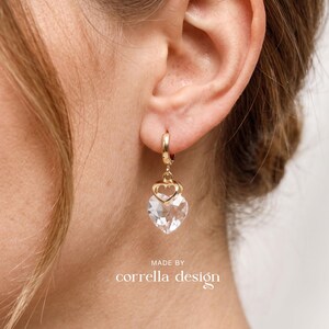 Könnte beinhalten: Ein goldener Creolen-Ohrring mit einem kleinen goldenen Herzanhänger und einem größeren, klaren, facettierten herzförmigen Kristallanhänger. Der Text "MADE BY corrella design" ist unten im Bild in Weiß unter dem Ohrring aufgedruckt.