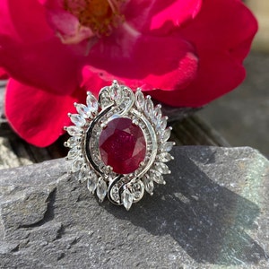 May include: A silver ring featuring a large, round, red ruby gemstone encircled by smaller, clear, marquise-shaped diamonds.