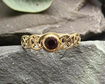 May include: A gold ring featuring a Celtic knot design and a dark red gemstone at its centre.
