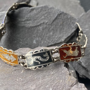 Puede incluir: Un brazalete de plata con tres dijes de esmalte. Los dijes están decorados con filigrana de plata y presentan un diseño de esmalte negro, naranja y rojo.