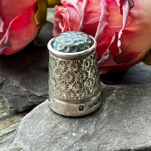 May include: A silver thimble featuring a repeating floral pattern across the body and a hammered, dimpled top. The base of the thimble displays small engraved hallmarks, including the number 4, indicating its metallic composition and origin.