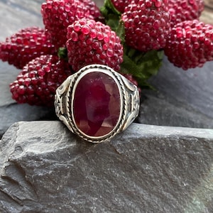 May include: A silver ring with an oval, faceted, deep red gemstone. The ring has an ornate, engraved design around the gemstone. The ring is displayed on a gray stone surface, with red berries in the background.
