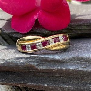 May include: A gold ring featuring a twisted design, adorned with alternating red gemstones and clear diamonds. The ring is presented on a dark grey stone surface, with a bright pink flower in the background.