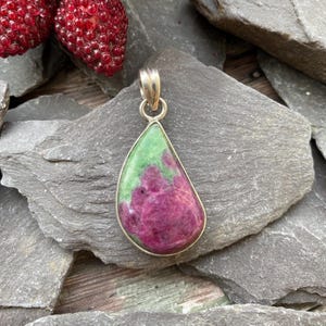 May include: A teardrop-shaped pendant with a silver-coloured bail. The pendant features a vibrant mix of colours, with a green top and a deep magenta lower section. The stone is set in a silver bezel. The pendant is displayed on a grey stone surface.