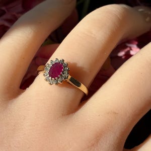 Pode incluir: Um anel de ouro com um rubi vermelho escuro de formato oval ao centro, rodeado por um halo de pequenas pedras brancas redondas e transparentes. A banda &eacute; de metal dourado polido e a crava&ccedil;&atilde;o apresenta uma disposi&ccedil;&atilde;o em forma de flor.