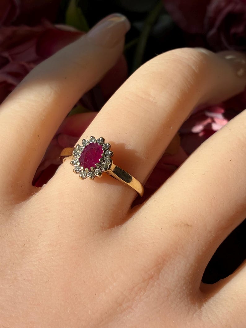 Pode incluir: Um anel de ouro com um rubi vermelho escuro de formato oval ao centro, rodeado por um halo de pequenas pedras brancas redondas e transparentes. A banda &eacute; de metal dourado polido e a crava&ccedil;&atilde;o apresenta uma disposi&ccedil;&atilde;o em forma de flor.
