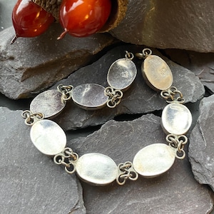 Pode incluir: Pulseira prateada com elos ovais ligados por conectores decorativos. A pulseira &eacute; apresentada sobre uma superf&iacute;cie cinza escura e texturizada, com bolotas no fundo. Os elos da pulseira t&ecirc;m uma apar&ecirc;ncia clara, possivelmente madrep&eacute;rola.