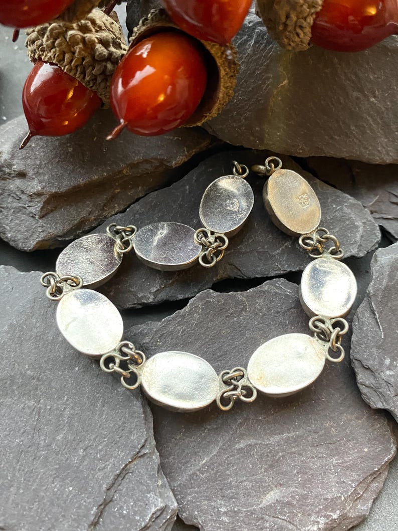Pode incluir: Pulseira prateada com elos ovais ligados por conectores decorativos. A pulseira &eacute; apresentada sobre uma superf&iacute;cie cinza escura e texturizada, com bolotas no fundo. Os elos da pulseira t&ecirc;m uma apar&ecirc;ncia clara, possivelmente madrep&eacute;rola.
