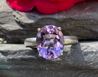 May include: A silver ring with a large, oval-shaped, light purple gemstone. The gemstone is faceted, catching the light and creating a sparkling effect. The ring is set against a natural, grey stone background, with a blurred red flower in the background.