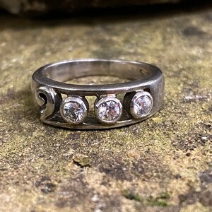 Peut inclure: Une bague en argent avec trois pierres rondes transparentes serties dans une bande incurvée. La bande présente un motif décoratif sur un côté.
