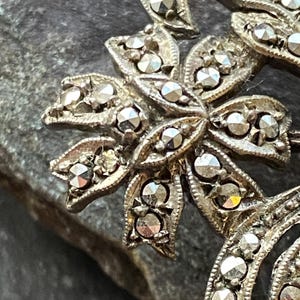 May include: Close-up of a vintage silver brooch with a floral design. The brooch features a central flower with multiple petals, each adorned with small, sparkling stones. The design includes curved, ribbon-like elements, also set with stones.