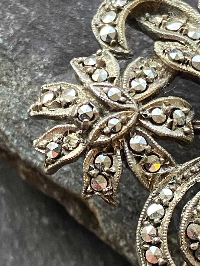 May include: Close-up of a vintage silver brooch with a floral design. The brooch features a central flower with multiple petals, each adorned with small, sparkling stones. The design includes curved, ribbon-like elements, also set with stones.
