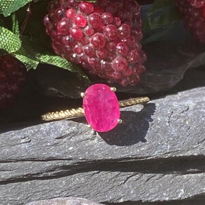 Könnte beinhalten: Ein goldener Ring mit einem leuchtend rosafarbenen ovalen Rubin. Der Ring hat ein gedrehtes Band.