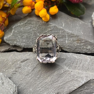Op de afbeelding: Een zilveren ring met een grote, rechthoekige, lichtroze edelsteen. De ring heeft een sierlijke, gedetailleerde zetting met een geschulpte rand. De edelsteen is facetgeslepen en vangt het licht. De ring ligt op een grijze stenen ondergrond.