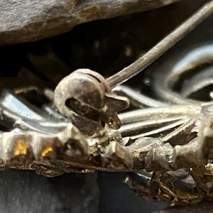 May include: Close-up of a silver brooch pin. The pin features an ornate design with a textured surface. The pin is set against a dark, textured background, highlighting its intricate details.