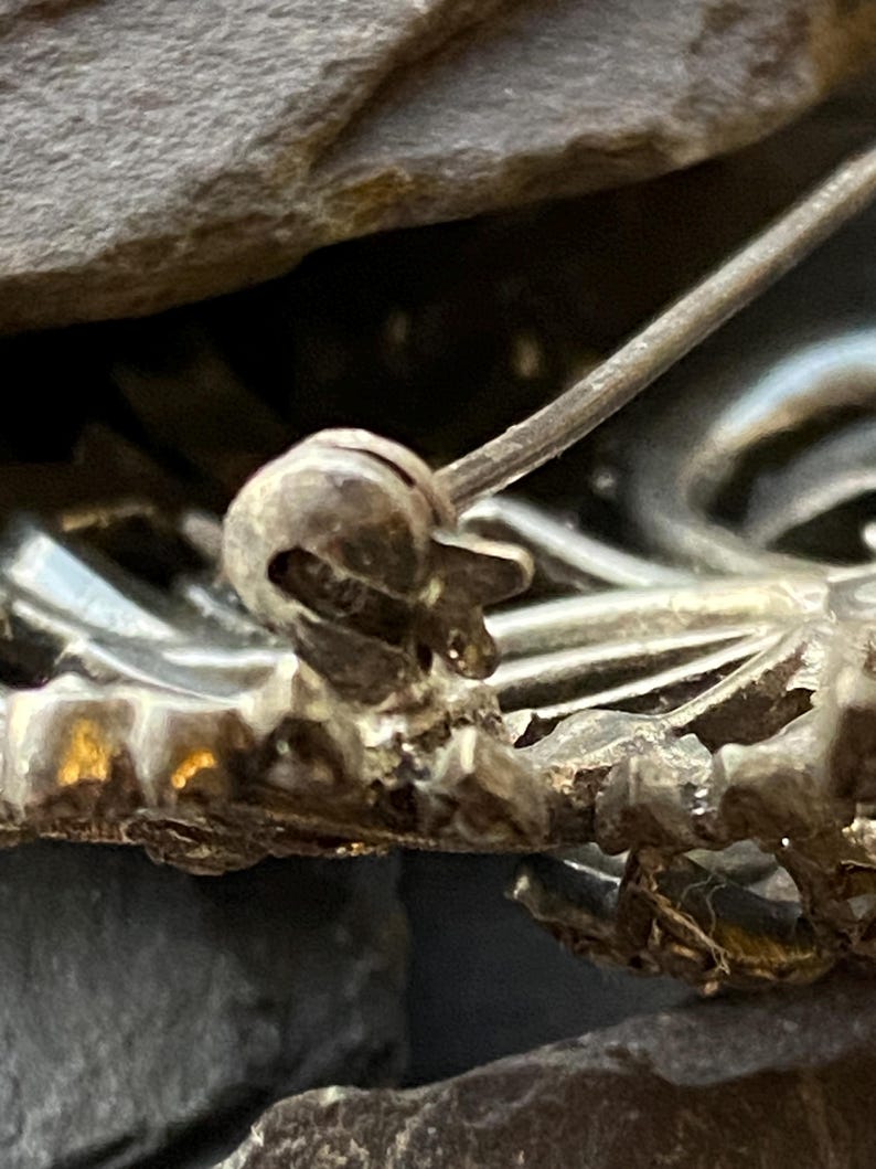May include: Close-up of a silver brooch pin. The pin features an ornate design with a textured surface. The pin is set against a dark, textured background, highlighting its intricate details.