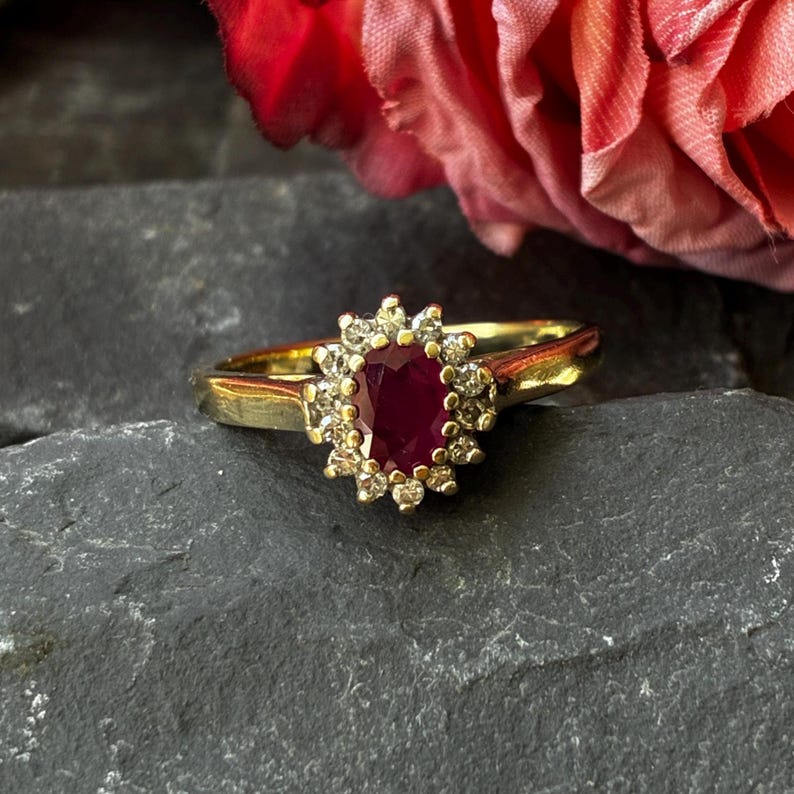 Pode incluir: Um anel de ouro com um rubi central de corte oval em vermelho profundo, rodeado por um halo de pequenos diamantes transparentes de corte redondo. A banda &eacute; de ouro amarelo polido e as pedras est&atilde;o dispostas num grupo central.