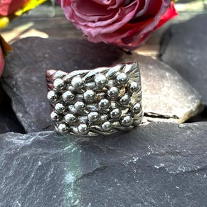 May include: A wide silver-toned ring featuring a textured surface covered in a grid of small, raised spherical beads. The band has a polished metallic finish with a repeating pattern of interconnected metal strands and rounded studs across the front.