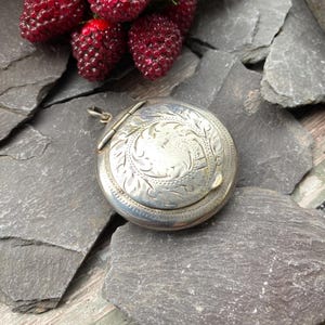 Op de afbeelding: Een ronde, zilveren locket met een gegraveerd bloemmotief op het deksel. De locket is open, waardoor de binnenkant zichtbaar is. De achtergrond bestaat uit donkergrijze leisteen en een tros rode bessen.