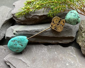 May include: A gold-coloured hat pin featuring an intricate filigree design and a turquoise stone. The pin is displayed on grey stones with green foliage, highlighting the jewellery's details and colour.
