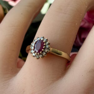 Pode incluir: Um anel de ouro com uma pedra central de rubi vermelho escuro de corte oval, rodeada por um halo de pequenas pedras de destaque brancas e claras. A banda &eacute; de metal de ouro amarelo polido com um design de crava&ccedil;&atilde;o em cacho.