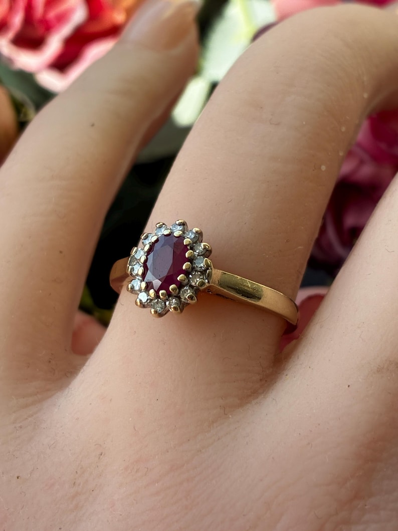 Pode incluir: Um anel de ouro com uma pedra central de rubi vermelho escuro de corte oval, rodeada por um halo de pequenas pedras de destaque brancas e claras. A banda &eacute; de metal de ouro amarelo polido com um design de crava&ccedil;&atilde;o em cacho.