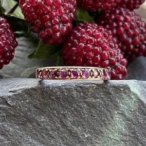 May include: A gold ring featuring a row of small, round, purple gemstones. The ring is displayed on a dark grey stone, with a background of ripe, red raspberries.