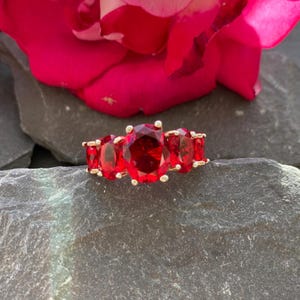 May include: A gold-toned ring featuring five oval-cut, vibrant red gemstones. The ring is set against a dark gray stone surface, with a blurred red rose in the background. The gemstones are the focal point of this elegant piece of jewelry.