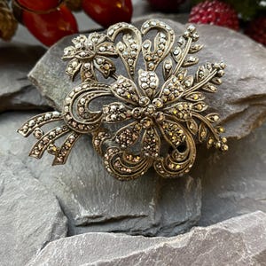 May include: Vintage silver-toned marcasite brooch with a floral design. The brooch features a central flower with surrounding leaves and swirling accents, all encrusted with small, sparkling marcasite stones. The brooch is set against a grey stone surface.
