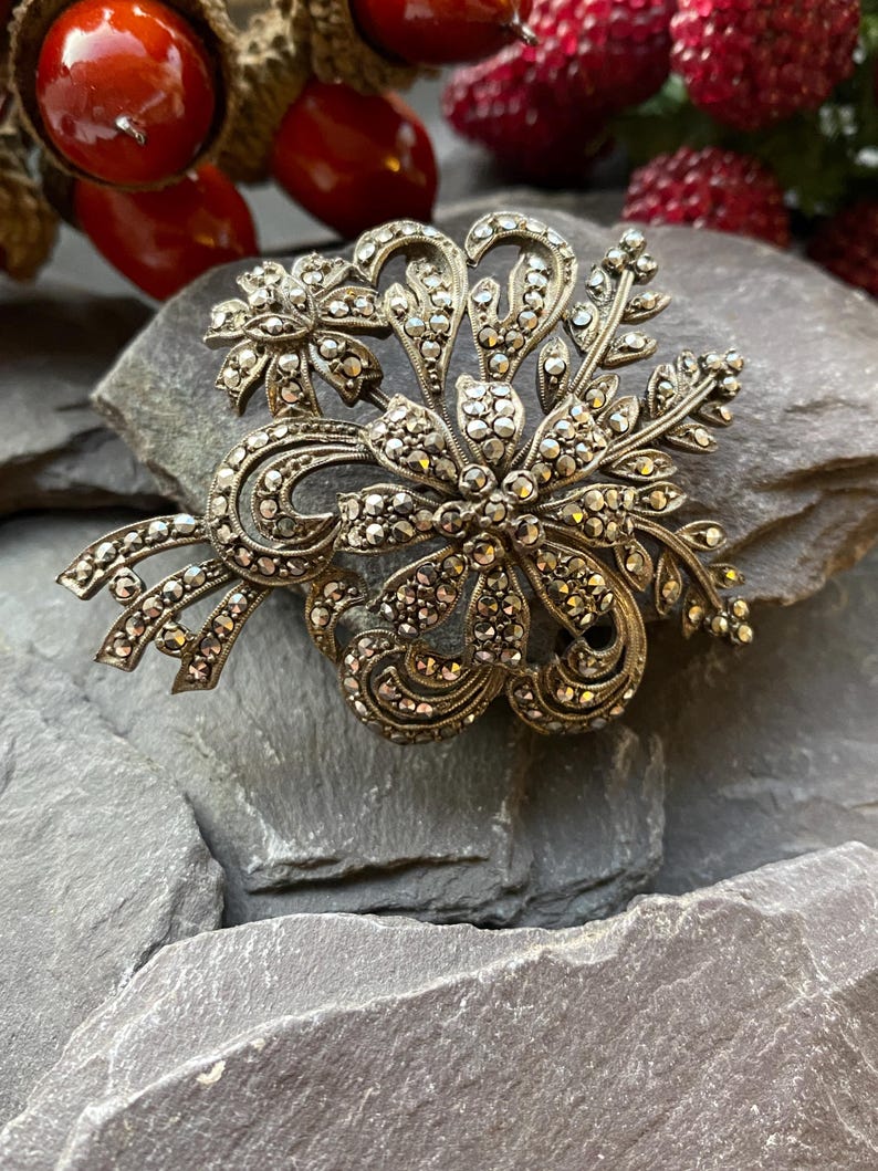 May include: Vintage silver-toned marcasite brooch with a floral design. The brooch features a central flower with surrounding leaves and swirling accents, all encrusted with small, sparkling marcasite stones. The brooch is set against a grey stone surface.