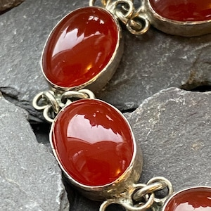 Pode incluir: Uma pulseira de prata com pedras preciosas ovais, polidas, vermelho-alaranjadas. As pedras s&atilde;o engastadas em uma moldura de prata e conectadas por elos de prata ornamentados. A pulseira &eacute; exibida em uma superf&iacute;cie escura e texturizada.