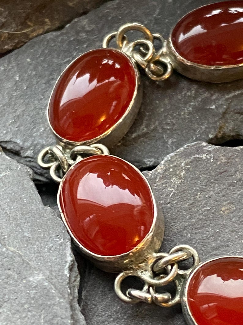 Pode incluir: Uma pulseira de prata com pedras preciosas ovais, polidas, vermelho-alaranjadas. As pedras s&atilde;o engastadas em uma moldura de prata e conectadas por elos de prata ornamentados. A pulseira &eacute; exibida em uma superf&iacute;cie escura e texturizada.