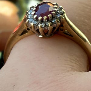 Pode incluir: Um anel de ouro com uma pedra preciosa de cor vermelho-escura de corte oval no centro, rodeada por um halo de pequenas pedras claras de corte redondo. O engaste &eacute; elevado e a banda &eacute; feita de metal de ouro amarelo polido e liso.