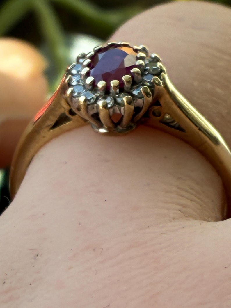 Pode incluir: Um anel de ouro com uma pedra preciosa de cor vermelho-escura de corte oval no centro, rodeada por um halo de pequenas pedras claras de corte redondo. O engaste &eacute; elevado e a banda &eacute; feita de metal de ouro amarelo polido e liso.