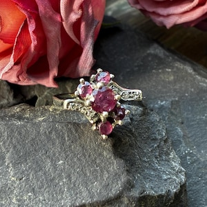 May include: A silver-toned ring featuring a cluster of round, deep pinkish-red gemstones set in a bypass-style band. The setting includes smaller, clear accent stones embedded along the metal shoulders, creating a layered, multi-stone design.