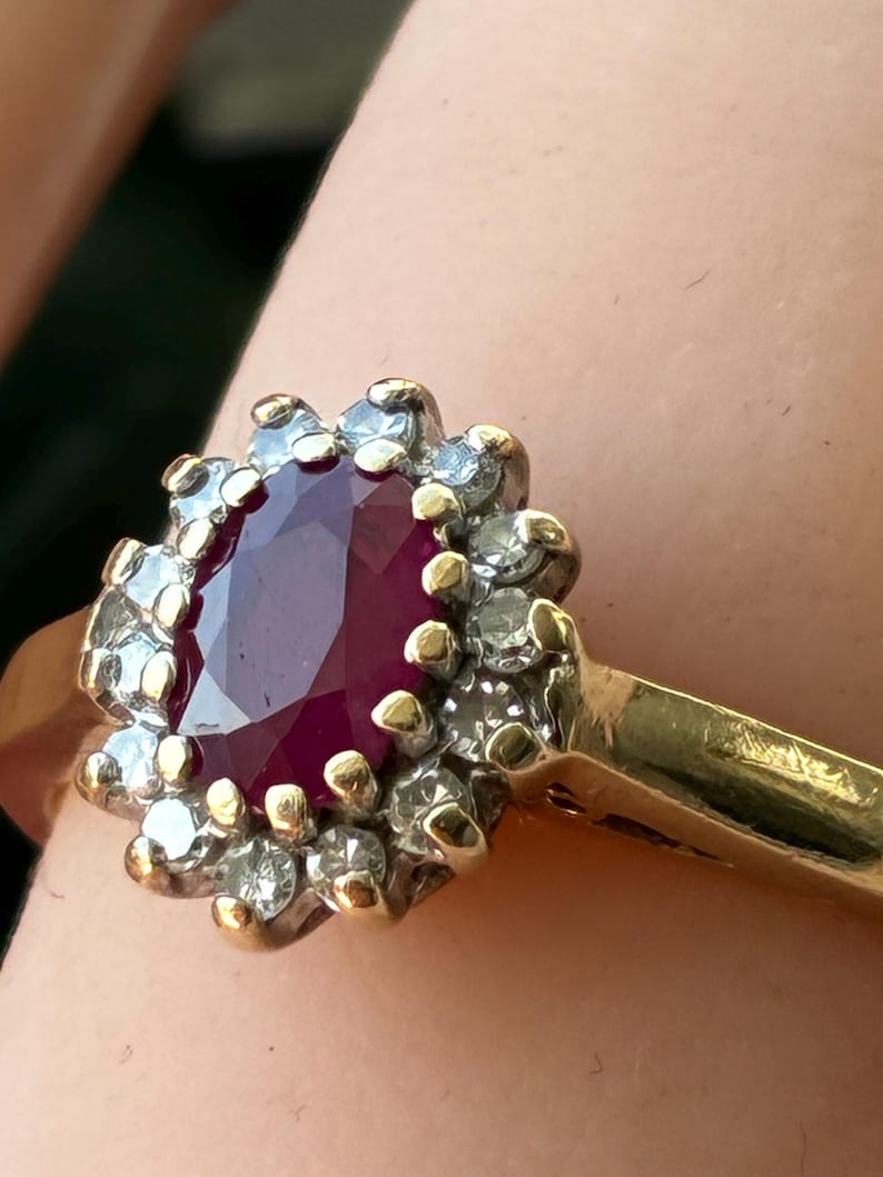Pode incluir: Um anel de ouro com um rubi vermelho profundo de corte oval no centro, rodeado por um halo de pequenos diamantes brancos claros e redondos cravados numa arma&ccedil;&atilde;o de tom prateado. A banda &eacute; feita de ouro amarelo polido.