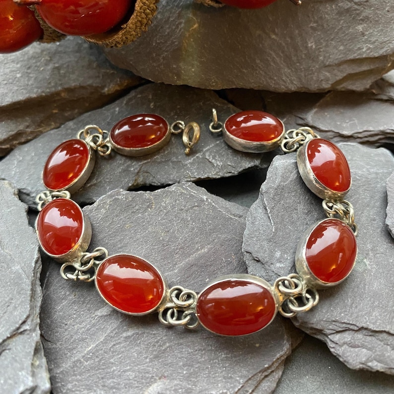Pode incluir: Uma pulseira de prata com pedras preciosas vermelhas ovais e polidas. As pedras s&atilde;o engastadas em bis&eacute;is individuais de prata e unidas com conectores decorativos de prata. A pulseira &eacute; exibida em uma superf&iacute;cie escura e texturizada.