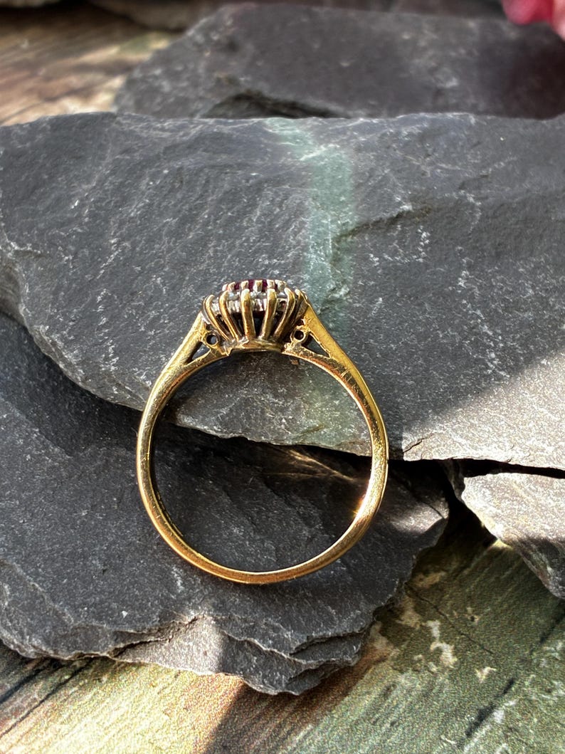 Pode incluir: Um anel de ouro com uma pedra preciosa vermelha escura central rodeada por um halo de pequenas pedras claras. A crava&ccedil;&atilde;o &eacute; elevada com uma galeria de garras e a banda &eacute; de metal dourado liso e polido, visto de perfil lateral.
